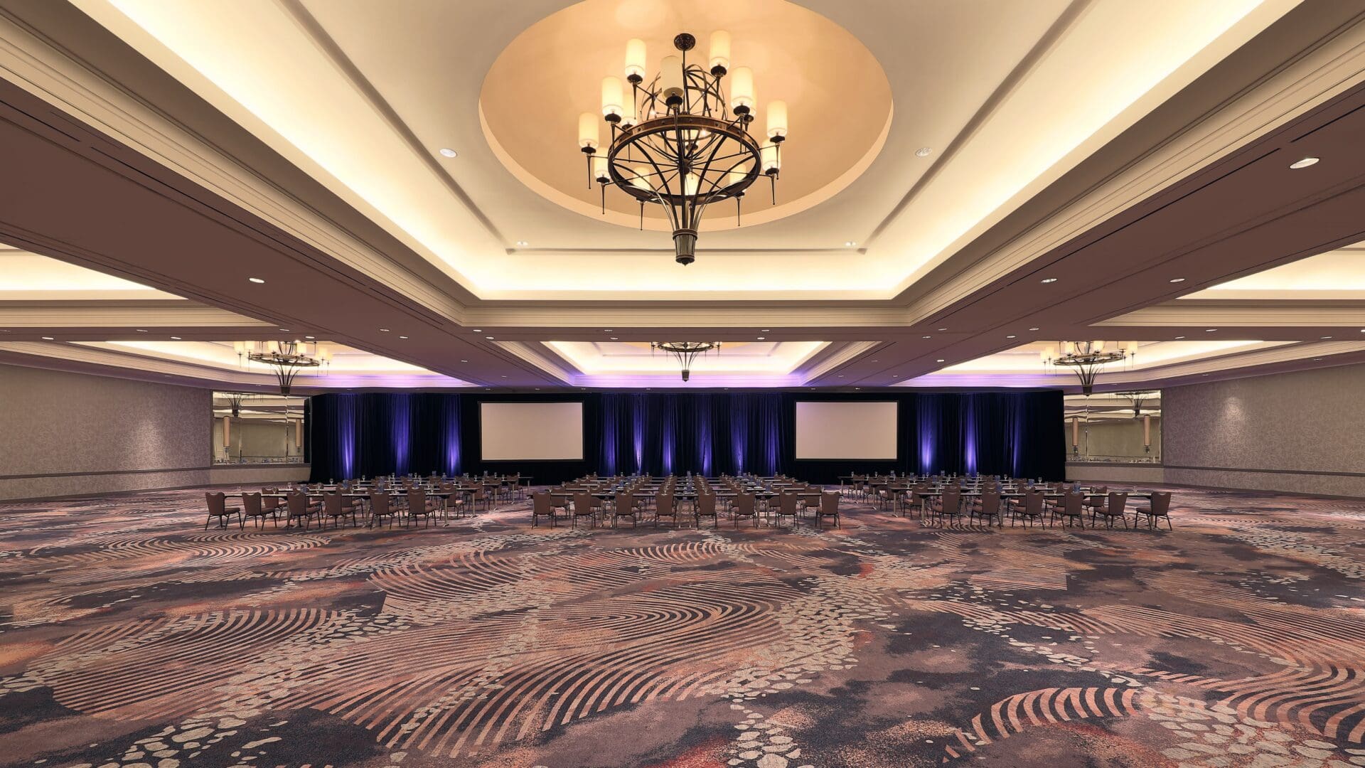 A large, elegant ballroom with a patterned carpet, chandeliers, and rows of chairs facing three projection screens and a stage area with blue lighting accents. The room is empty and ready for an event or conference.