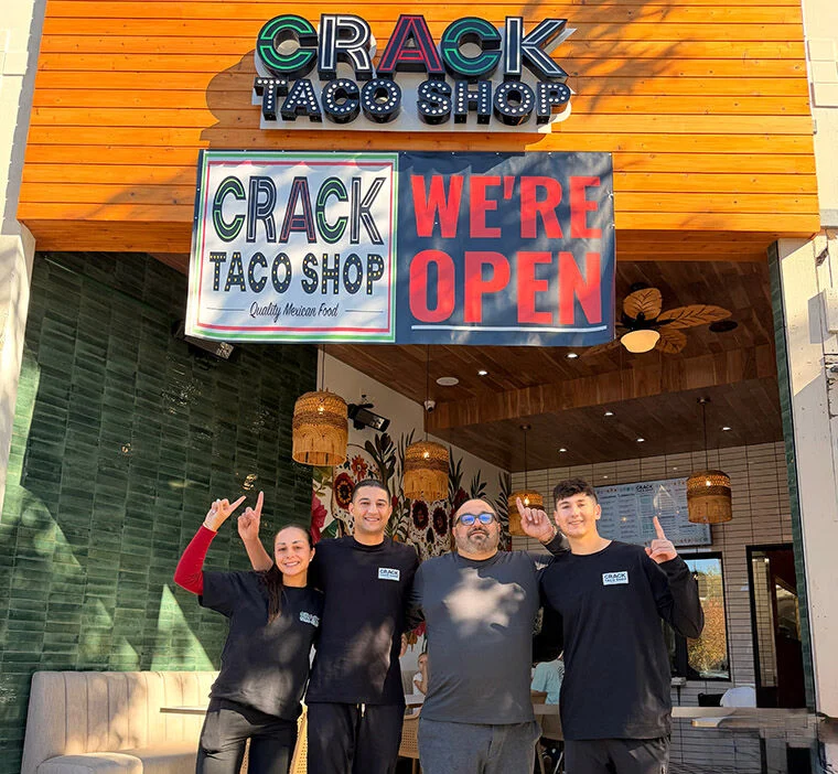 Four people stand smiling outside Crack Taco Shop under signs that say “WE’RE OPEN” and “Quality Mexican Food.” They gesture with peace signs and point upward, celebrating the shop’s opening.