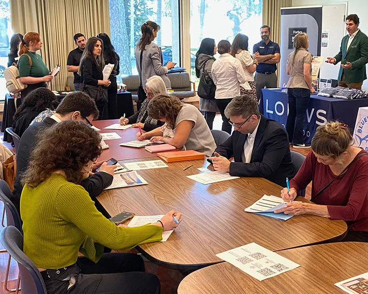 People sit at a table filling out forms at a job fair event, while others stand and speak with recruiters at booths in the background. The atmosphere is busy and focused.