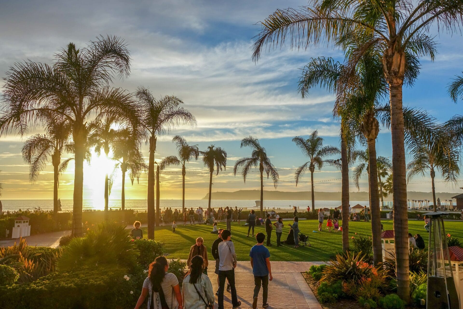 People walk and gather in a grassy park lined with tall palm trees at sunset, with the ocean and distant mountains in the background under a partly cloudy sky.
