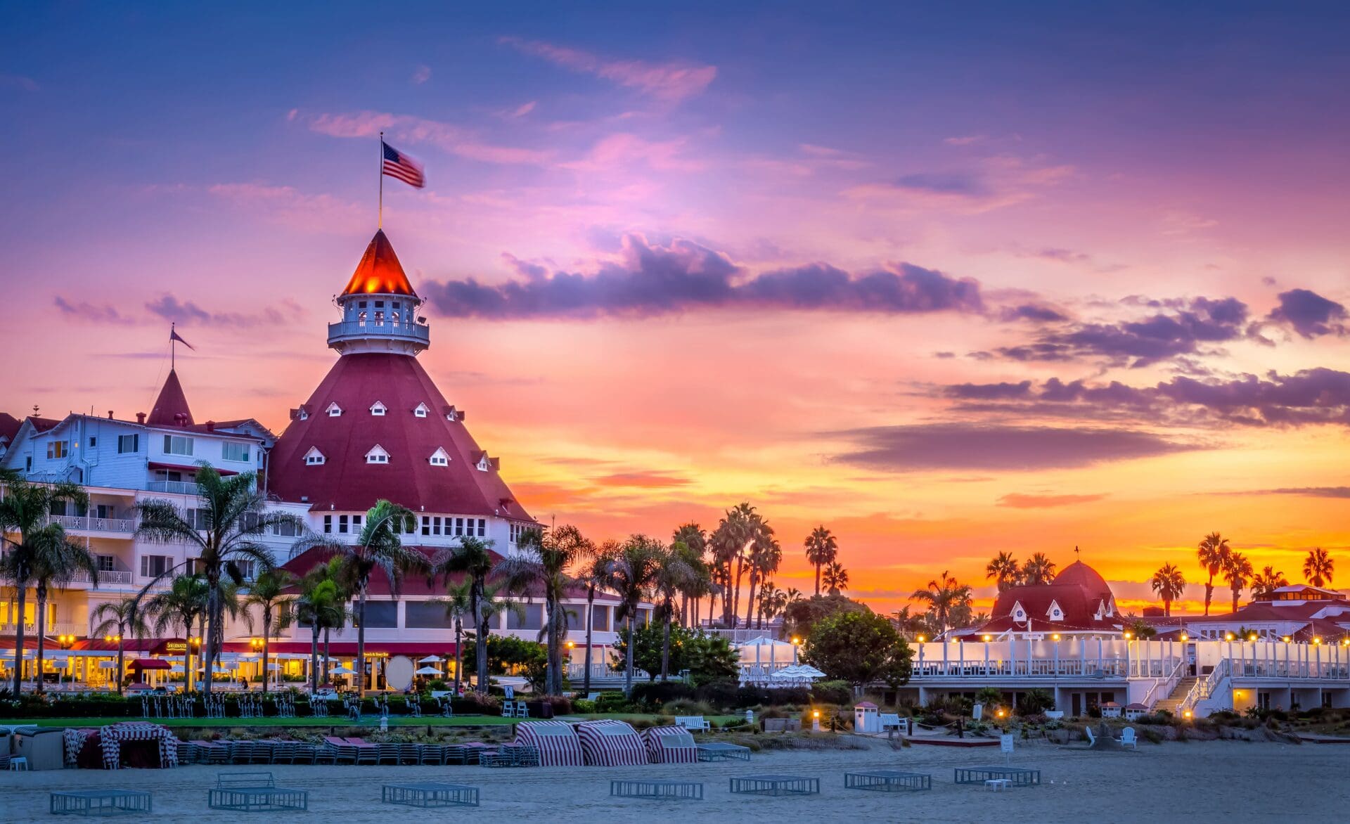 A historic beachfront hotel with a red turret roof stands among palm trees at sunset, with a vibrant sky of purple, orange, and pink hues and the American flag flying above.