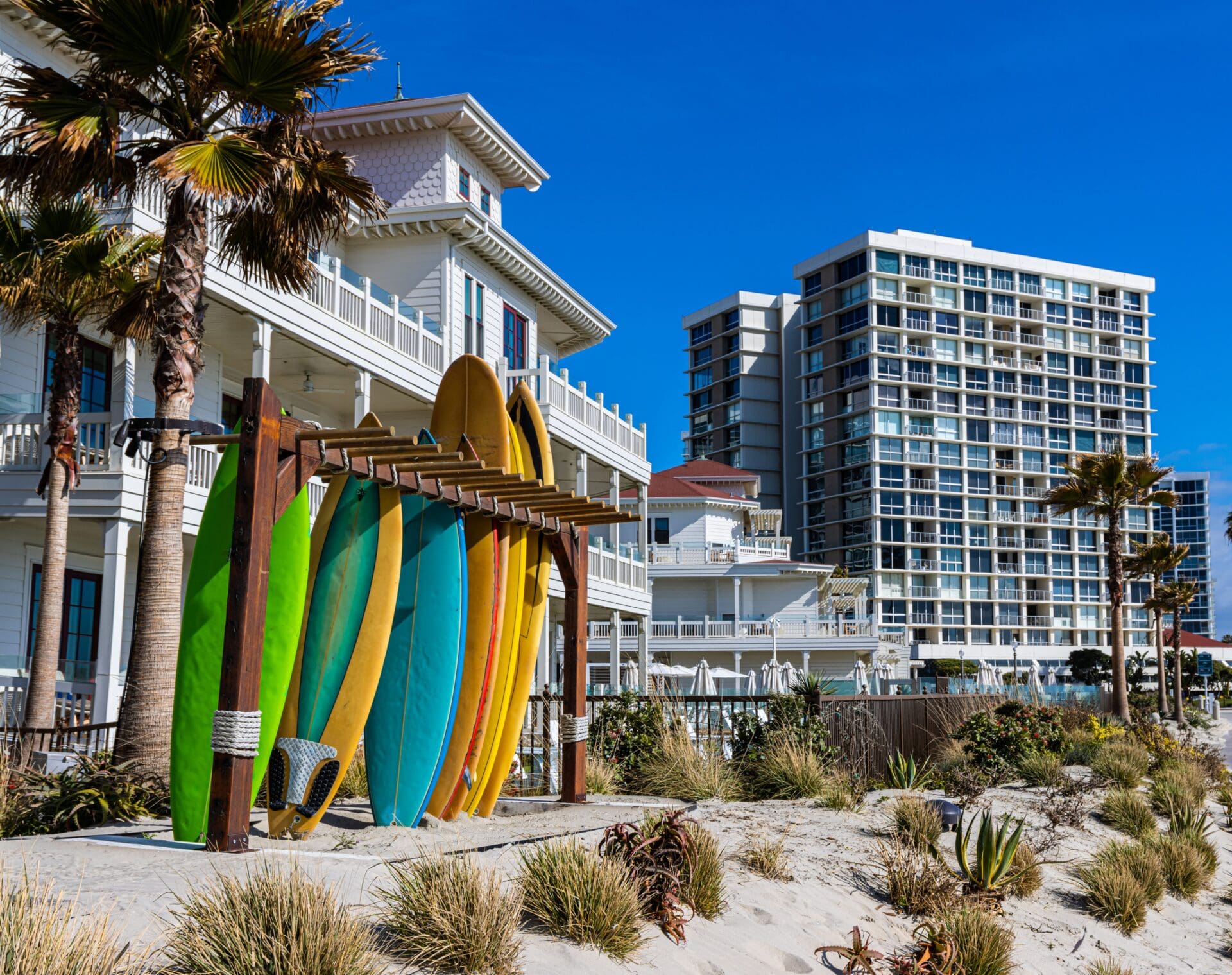 Colorful surfboards are propped against a wooden rack on a sandy beach, with palm trees and modern beachfront buildings in the background under a clear blue sky.