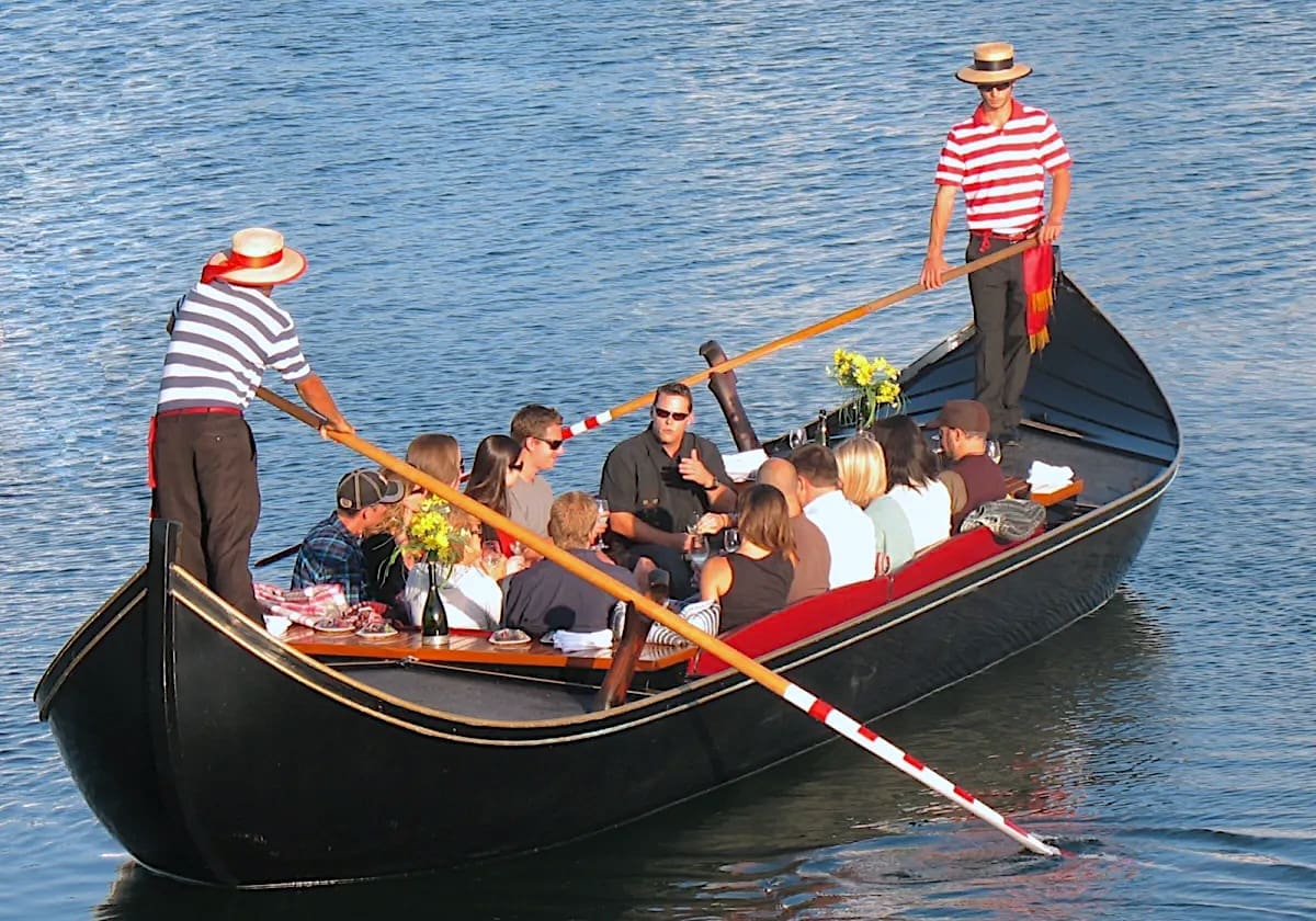 A group of people sit in a black gondola decorated with flowers, enjoying a ride on calm water, while two gondoliers in striped shirts and hats steer the boat with long oars.