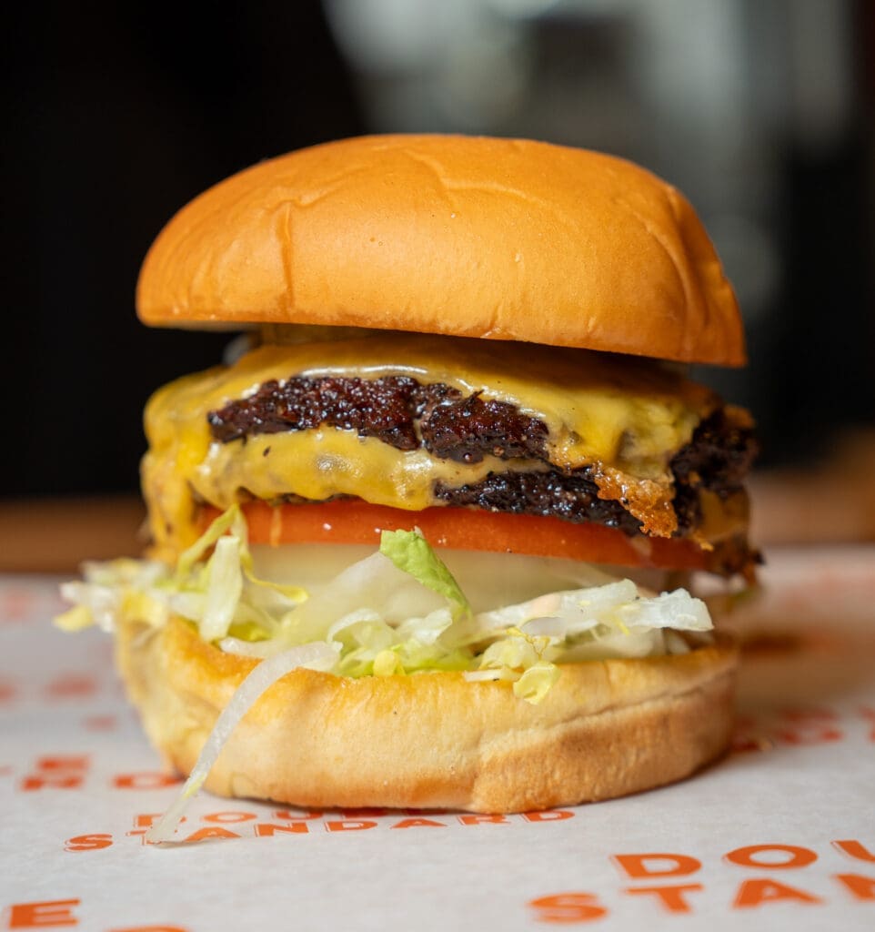 A close-up of a cheeseburger with two beef patties, melted cheese, tomato slices, onions, and shredded lettuce in a soft, golden bun, placed on a paper wrapper.