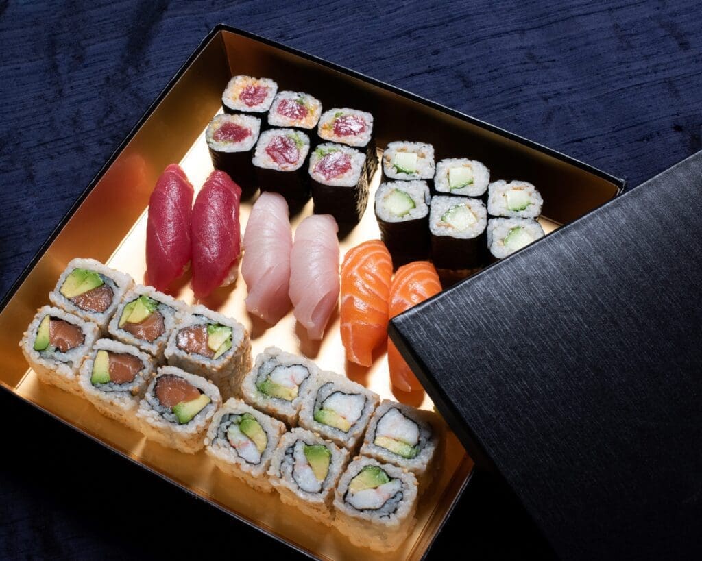 An opened black box filled with assorted sushi, including tuna, salmon, and yellowtail nigiri, maki rolls with seaweed, and inside-out rolls with rice, avocado, and fish, set on a dark blue surface.