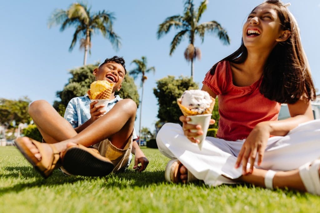 Two children sit on grass under palm trees, laughing and enjoying ice cream cones on a sunny day.