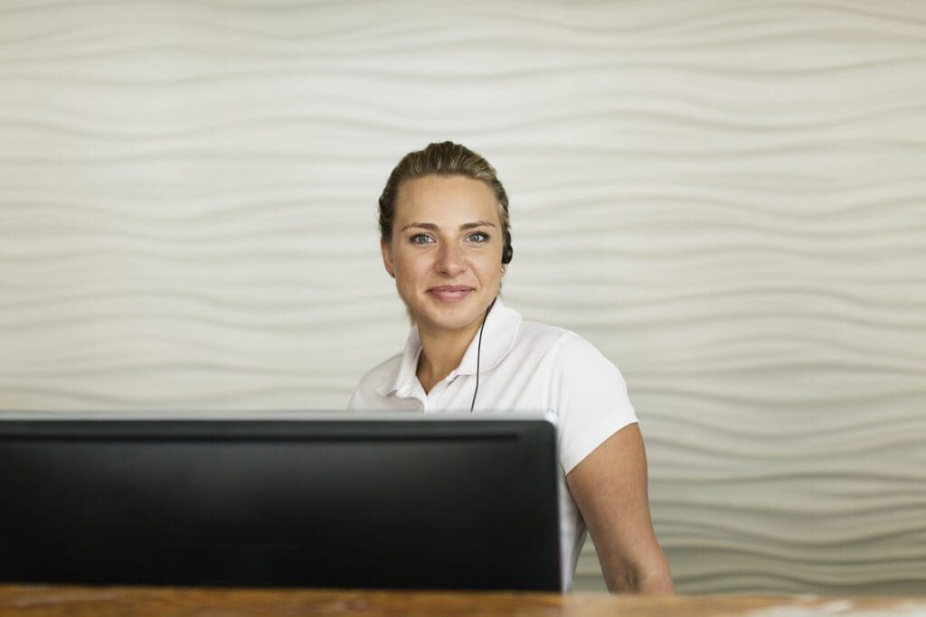 A woman wearing a white shirt and headset sits behind a computer at a reception desk, smiling. The background features a white wall with a wavy textured pattern.