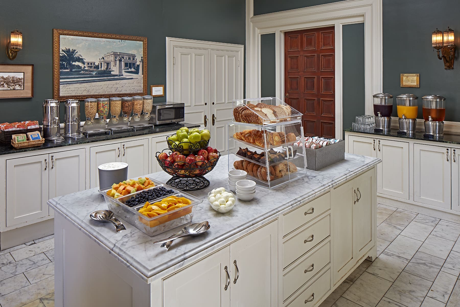 A breakfast buffet features cereals, pastries, bread, fruit, juice, eggs, and coffee, all arranged on white cabinets and a marble island in a well-lit, elegant room.