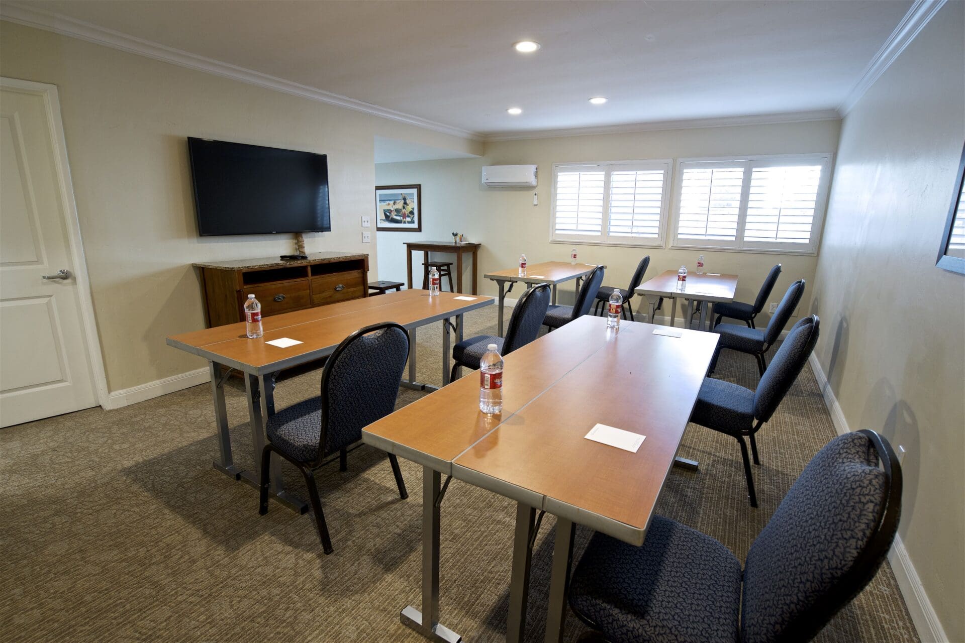 Small conference room with three rectangular tables, black chairs, bottled water, and notepads. A large TV is mounted on the wall, and windows with white shutters let in light. The room has beige walls and carpet.