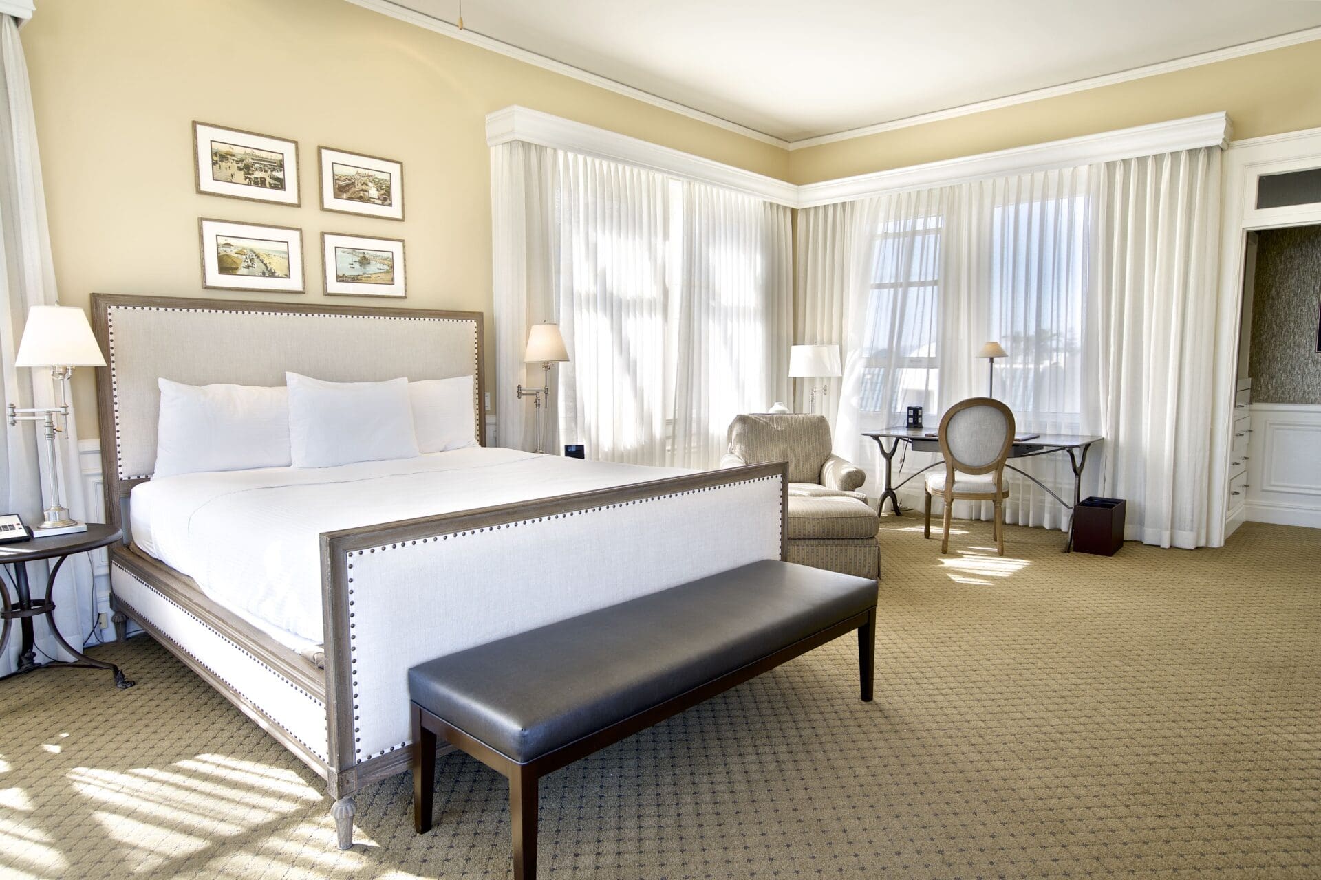 Bright hotel room with a large bed, cushioned bench, armchair, desk, and chair. Light streams in through sheer curtains covering multiple windows, and framed art hangs above the bed. The decor is neutral and elegant.