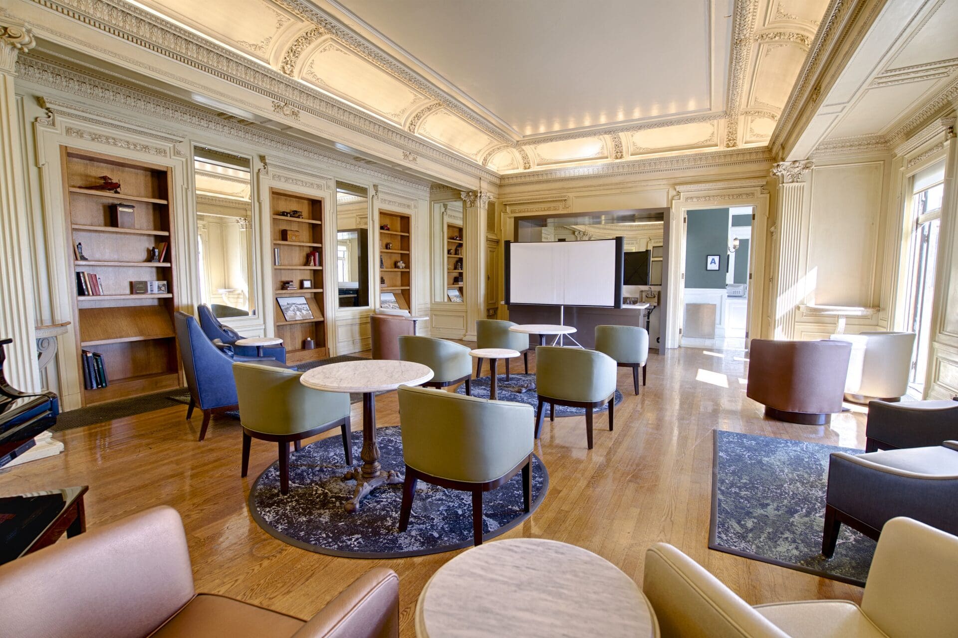 A spacious, elegant lounge with high ornate ceilings, round tables, modern chairs, bookshelves, and large windows. A whiteboard stands at the center, and natural light fills the room.