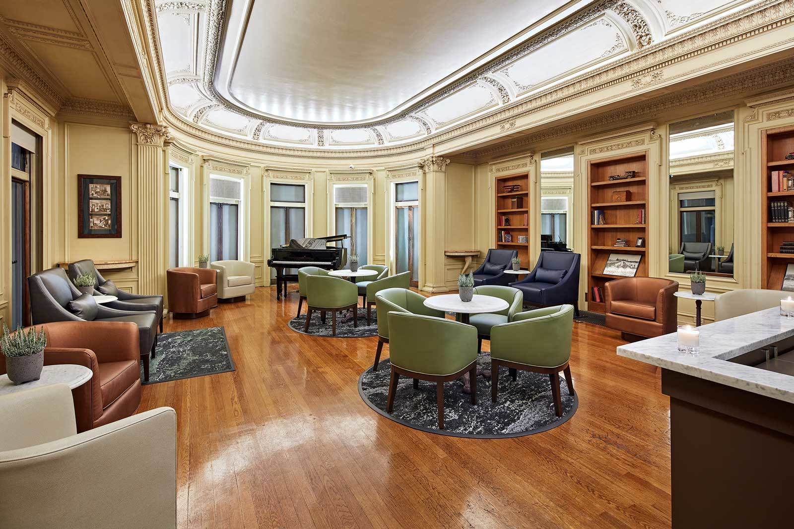 Elegant lounge with round tables, green and brown chairs, hardwood floors, a grand piano, bookshelves, and ornate ceiling details. Large windows let in natural light, creating a warm, inviting atmosphere.