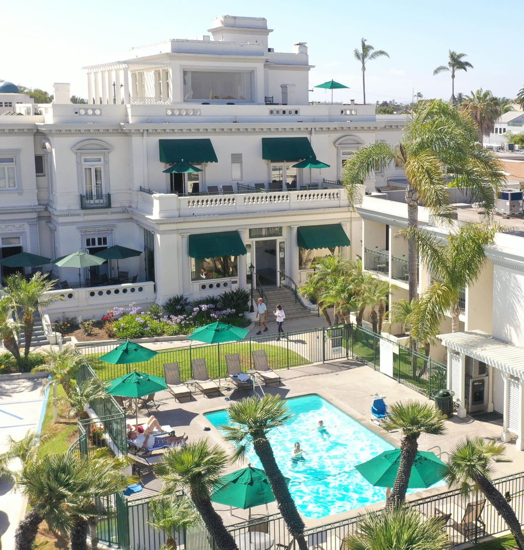 Aerial view of a white mansion with green awnings, a fenced outdoor swimming pool, palm trees, lounge chairs, umbrellas, and several people relaxing and swimming on a sunny day.