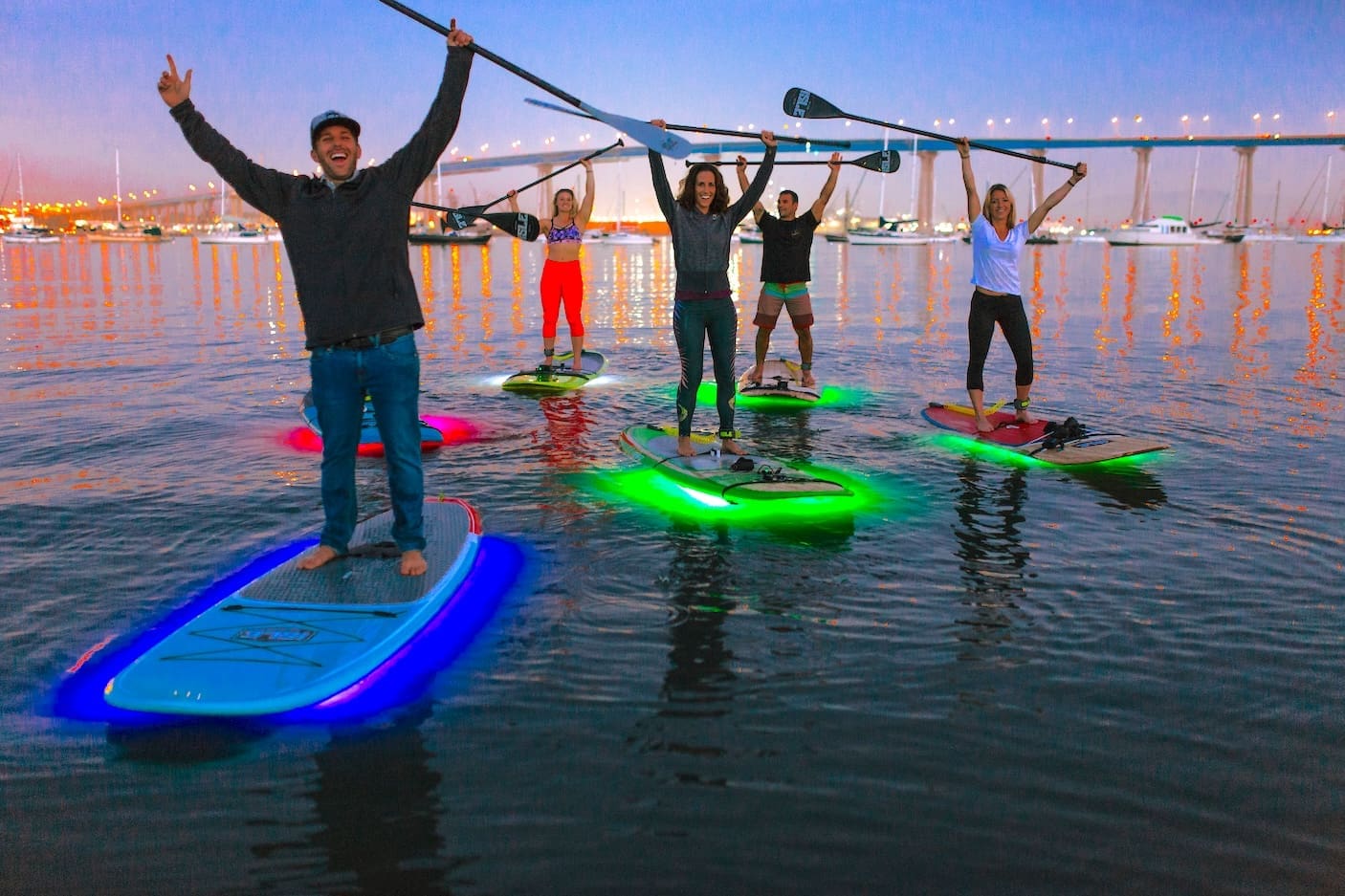 Five people on paddleboards with glowing lights stand on calm water at dusk, holding paddles above their heads, smiling, with a bridge and boats visible in the background.