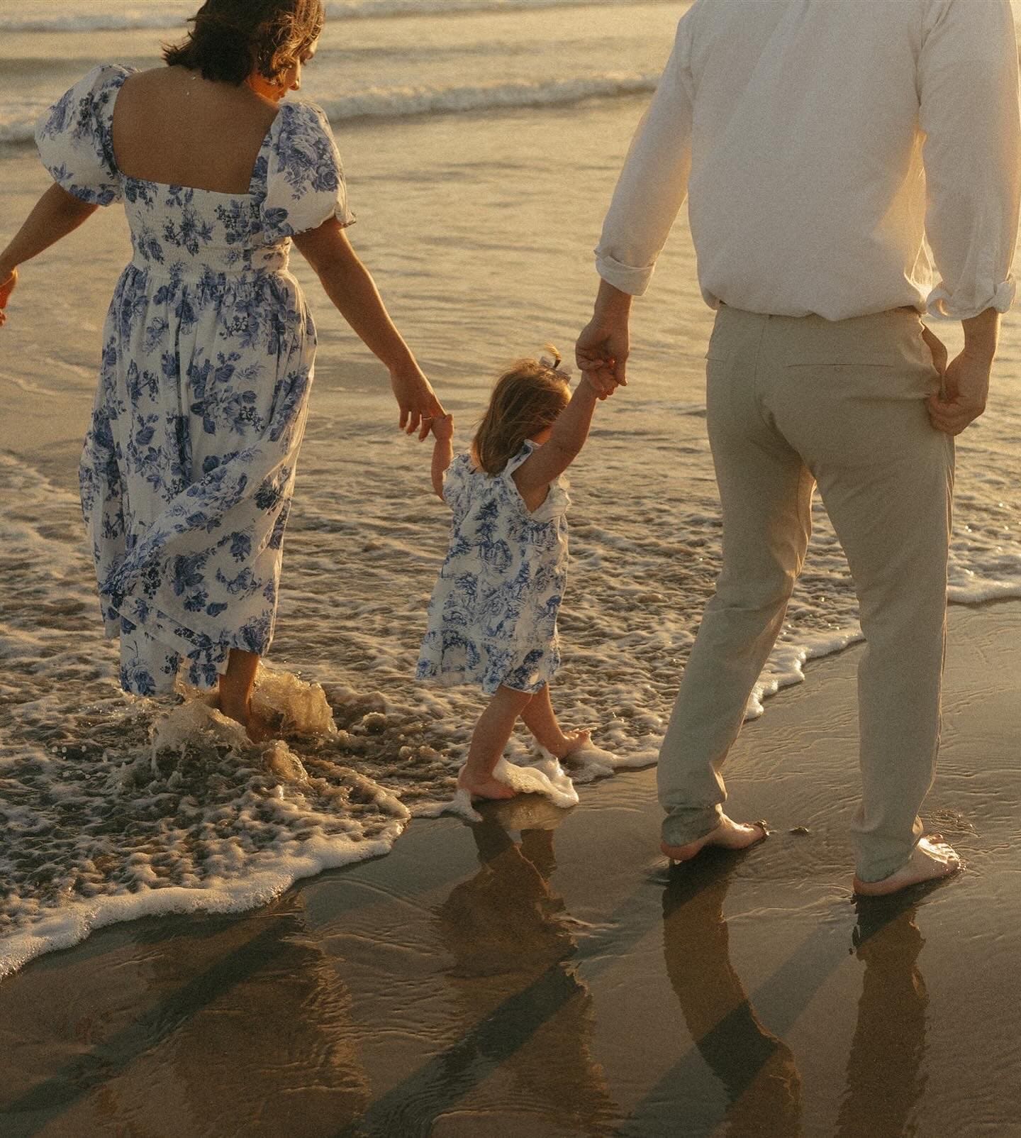 A family walks barefoot along the beach at sunset. A child in a blue and white dress holds hands with two adults, who are also dressed in light colors, as they walk through shallow waves.