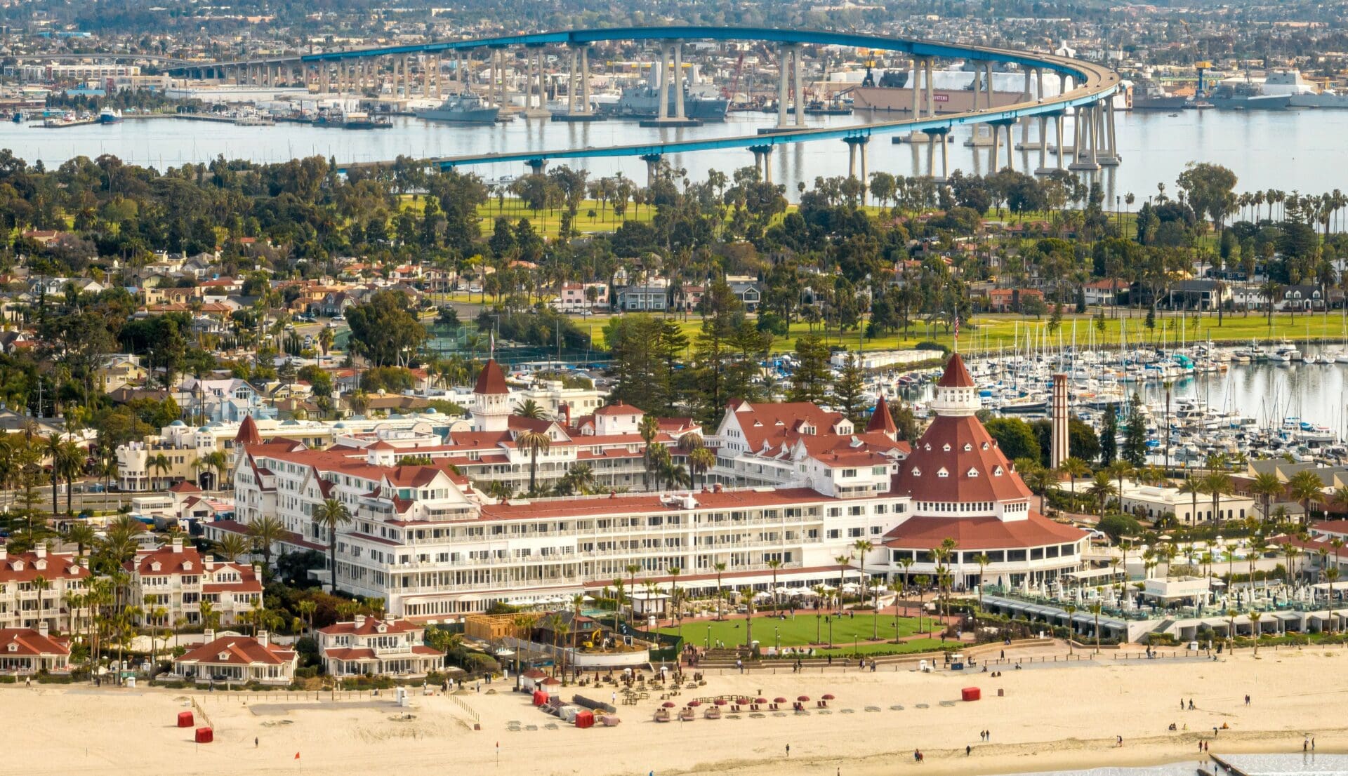 Aerial view of the historic Hotel del Coronado with its red roofs, sandy beach, and palm trees, and the blue Coronado Bridge and marina in the background in San Diego, California.