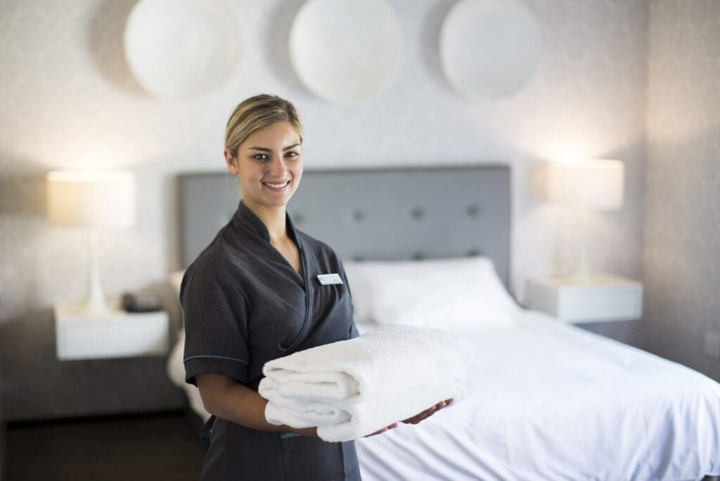 A hotel housekeeper in uniform stands smiling, holding neatly folded white towels in front of a well-made bed in a modern, tidy hotel room with two bedside lamps and decorative wall plates.