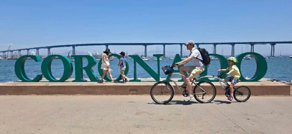 A man and a child ride bicycles past large “CORONADO” letters by the water, with a bridge and people walking in the background under a sunny sky.