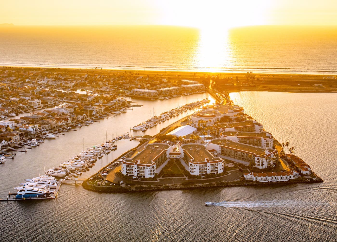 Aerial view of a triangular marina at sunset, with boats docked along the edges, modern buildings, and a speedboat moving through the water, as the sun sets over the ocean in the background.