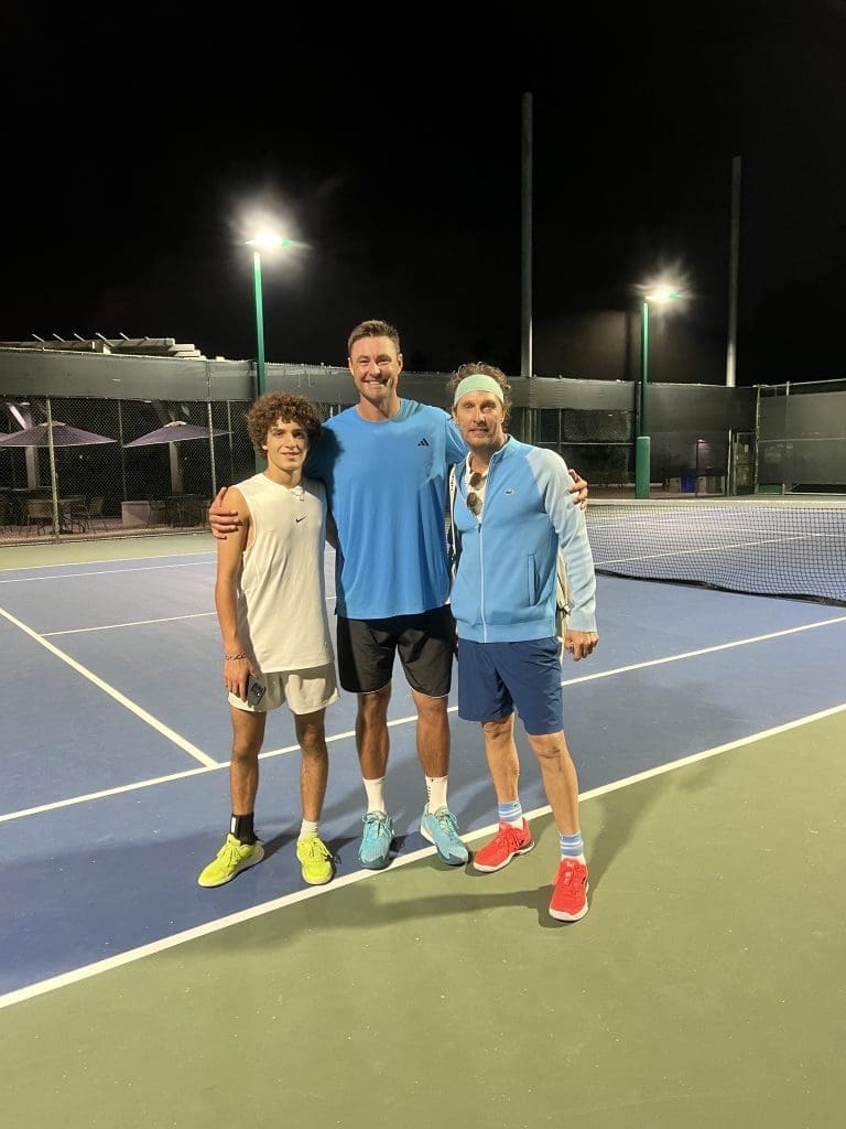 Three men stand together on a tennis court at night, smiling for the camera. One is in a white outfit, another in a blue t-shirt and shorts, and the third in a blue tracksuit and headband. The court is lit by overhead lights.