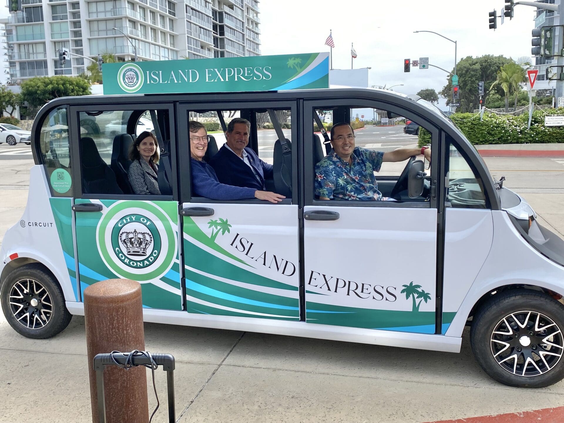 Four people sit smiling inside an “Island Express” electric shuttle with city and palm tree graphics, parked on a city street near a modern building and traffic signals. The driver wears a floral shirt.