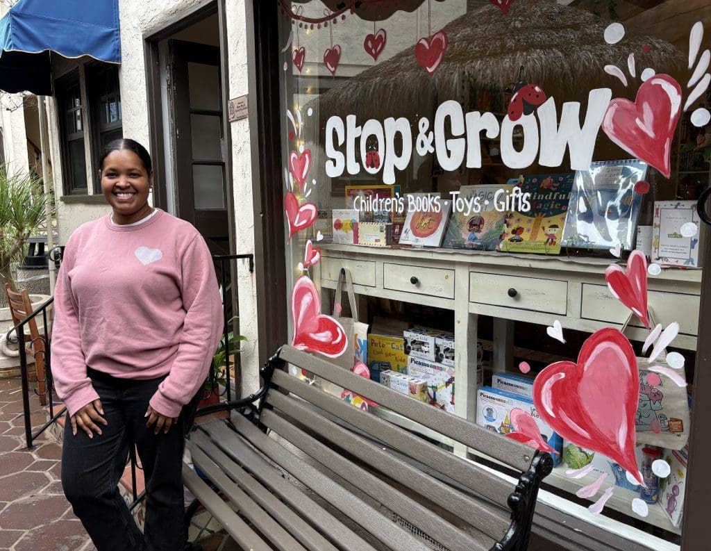 A person in a pink sweater stands smiling next to a bench outside a shop called “Stop & Grow,” decorated with painted red hearts; the window displays children’s books, toys, and gifts.