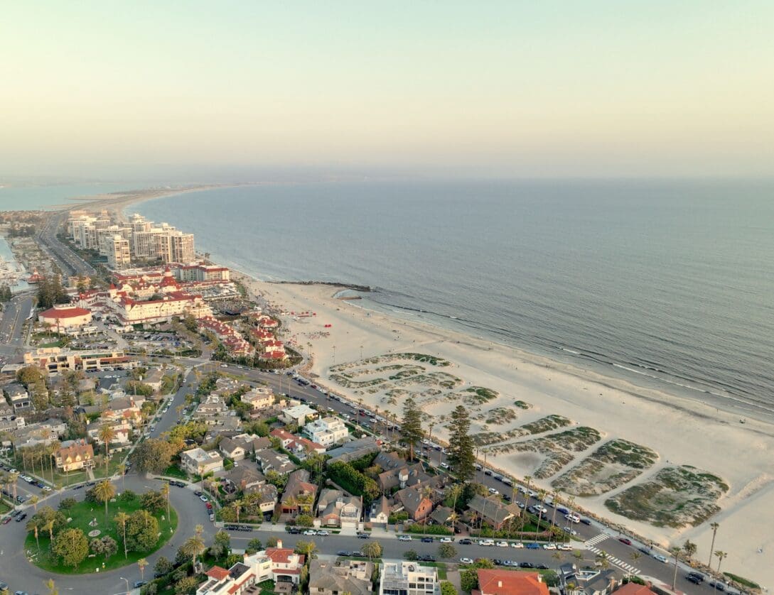 Aerial view of a coastal city with sandy beaches, ocean waves, and shoreline development. Residential neighborhoods and tall buildings border the beach, with roads and greenery visible in the foreground.