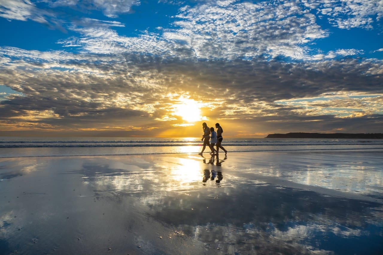 Three people walk along a reflective beach at sunset, silhouetted against a dramatic sky filled with clouds and golden sunlight. The ocean and distant land are visible in the background.