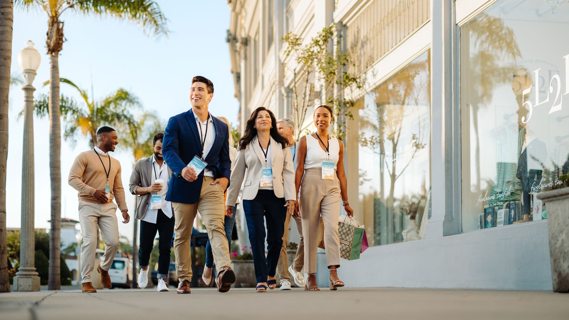 A group of professionally dressed people walk together on a sunny sidewalk lined with palm trees and storefronts, smiling and wearing conference badges.