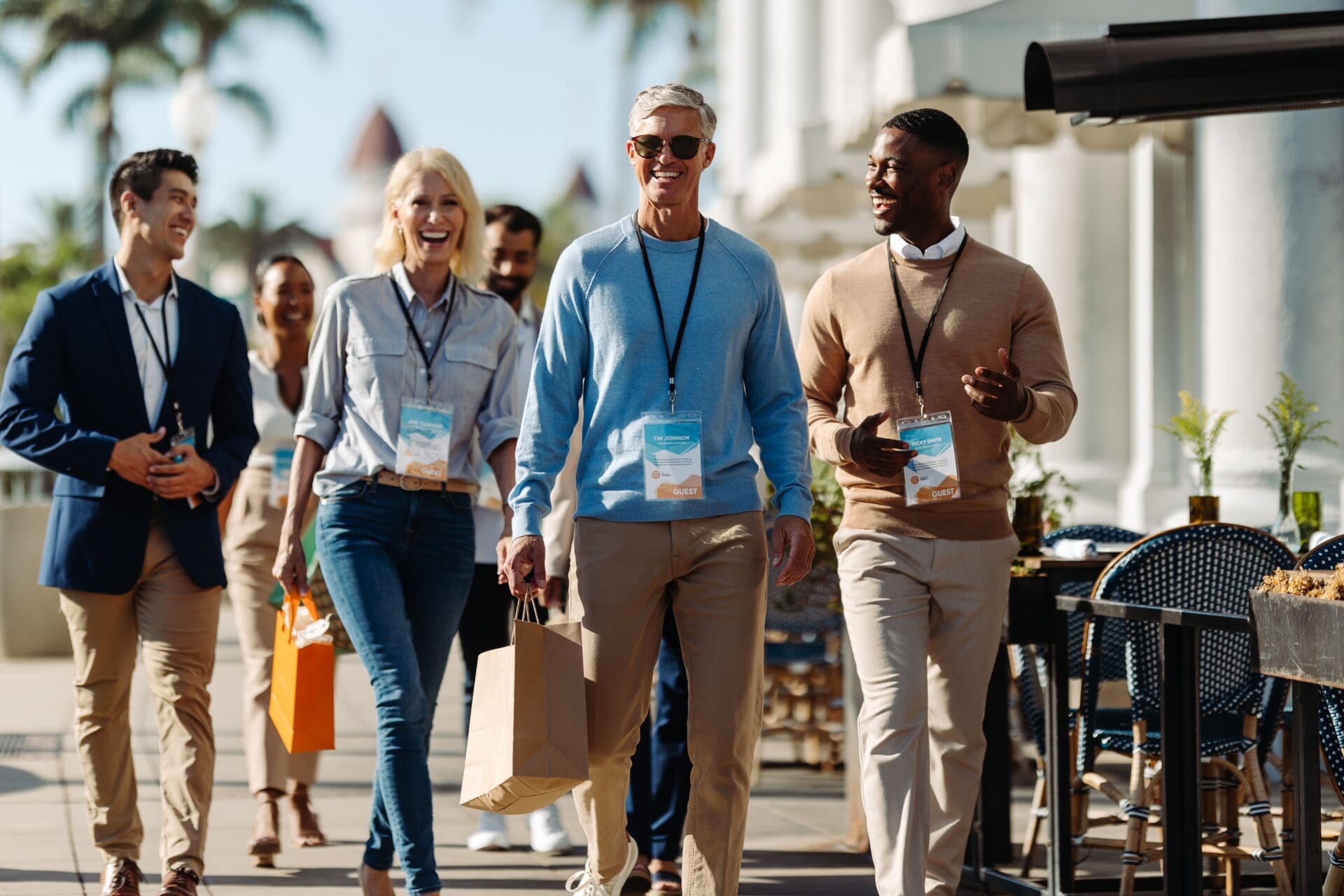A group of professionally dressed people, wearing conference badges, smile and walk outdoors together on a sunny day. Some are carrying shopping bags. Palm trees and a building are visible in the background.