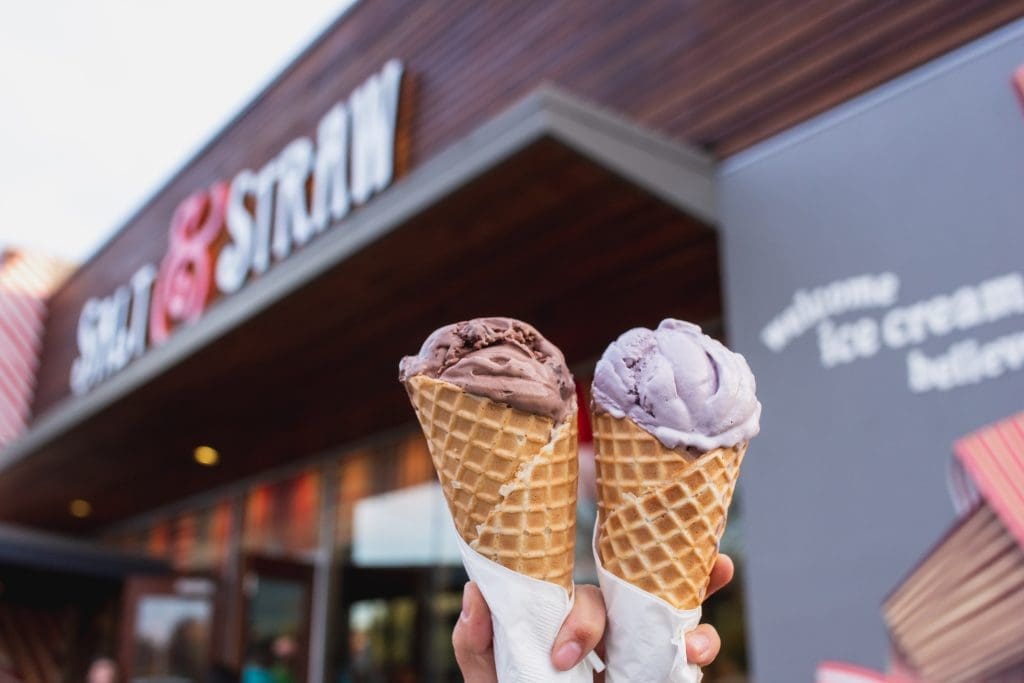 Two hands hold waffle cones with scoops of chocolate and lavender ice cream in front of a Salt & Straw ice cream shop.