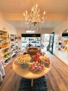A boutique shop interior with a round table displaying colorful gifts and plush toys under a crystal chandelier. Shelves line the walls holding various products, and the glass door is open to a street with parked cars outside.