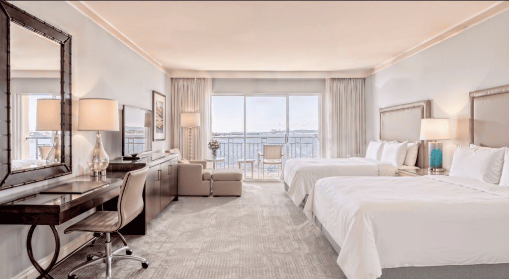 Spacious hotel room with two large beds, a desk, armchair, lamps, and a large window with a view of water and boats outside. Neutral tones and modern furnishings create a bright, relaxing atmosphere.