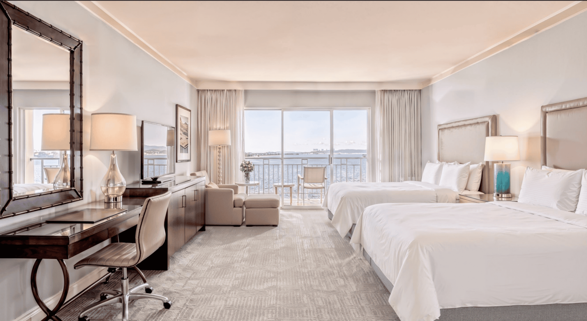 Spacious hotel room with two large beds, a desk, armchair, lamps, and a large window with a view of water and boats outside. Neutral tones and modern furnishings create a bright, relaxing atmosphere.