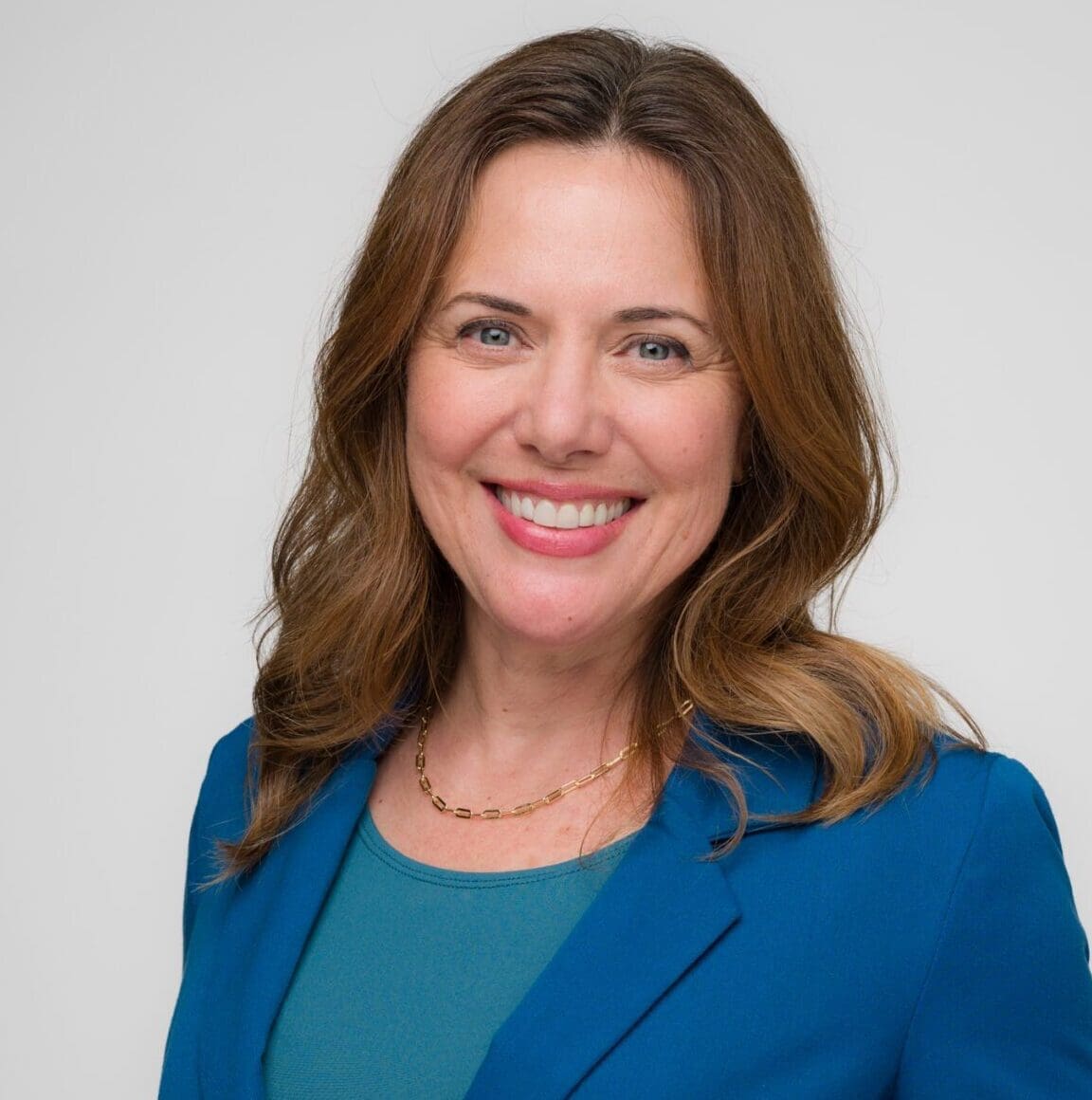 A woman with wavy brown hair smiles at the camera. She is wearing a teal top, a blue blazer, and a gold necklace. The background is plain and light-colored.