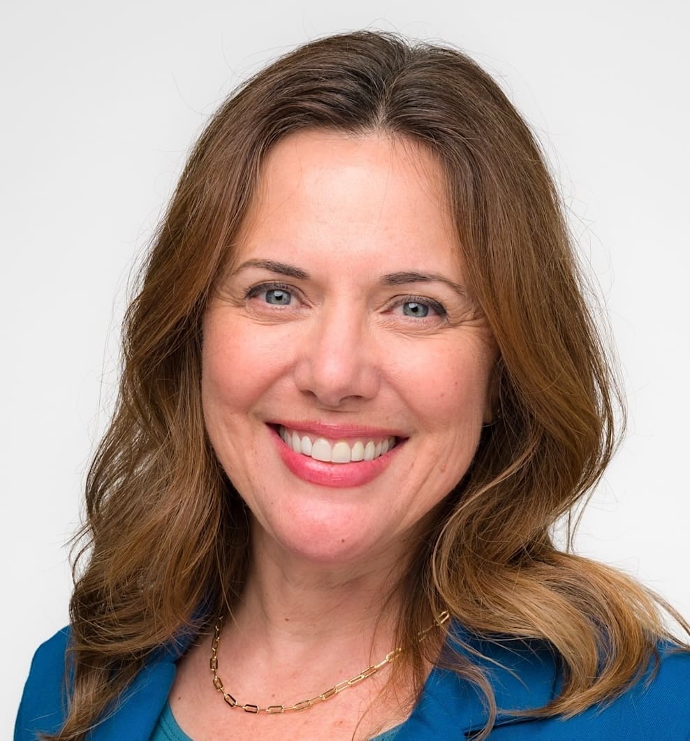 A woman with long brown hair smiles at the camera. She is wearing a blue blazer and a gold necklace, with a light background.