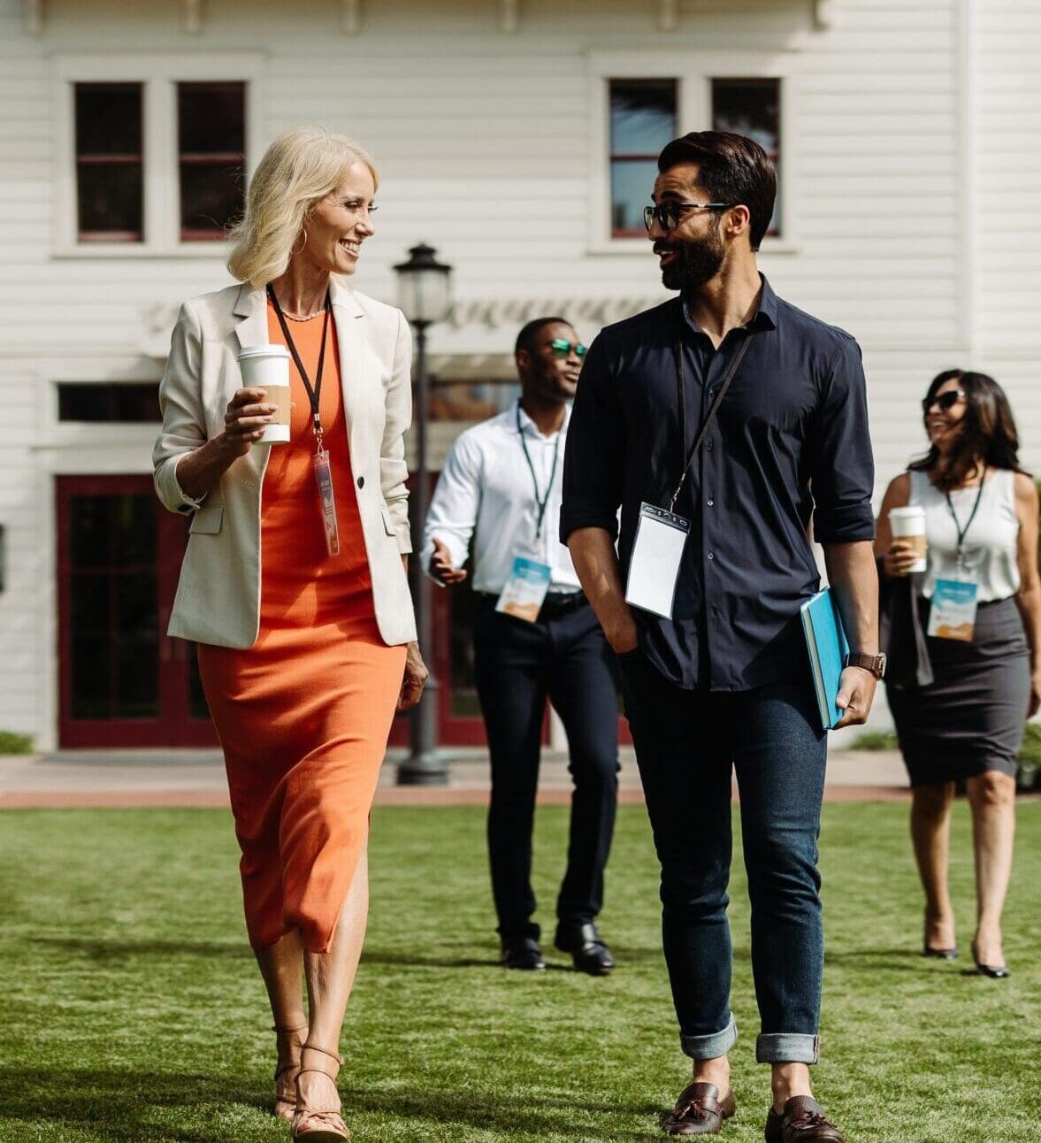 A group of professionally dressed people walk and talk outside on a lawn, wearing conference name badges, with two people in front smiling and holding coffee cups and folders. A white building is in the background.