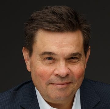 A middle-aged man with short brown hair, wearing a dark pinstripe suit and white shirt, smiling slightly against a plain dark background.