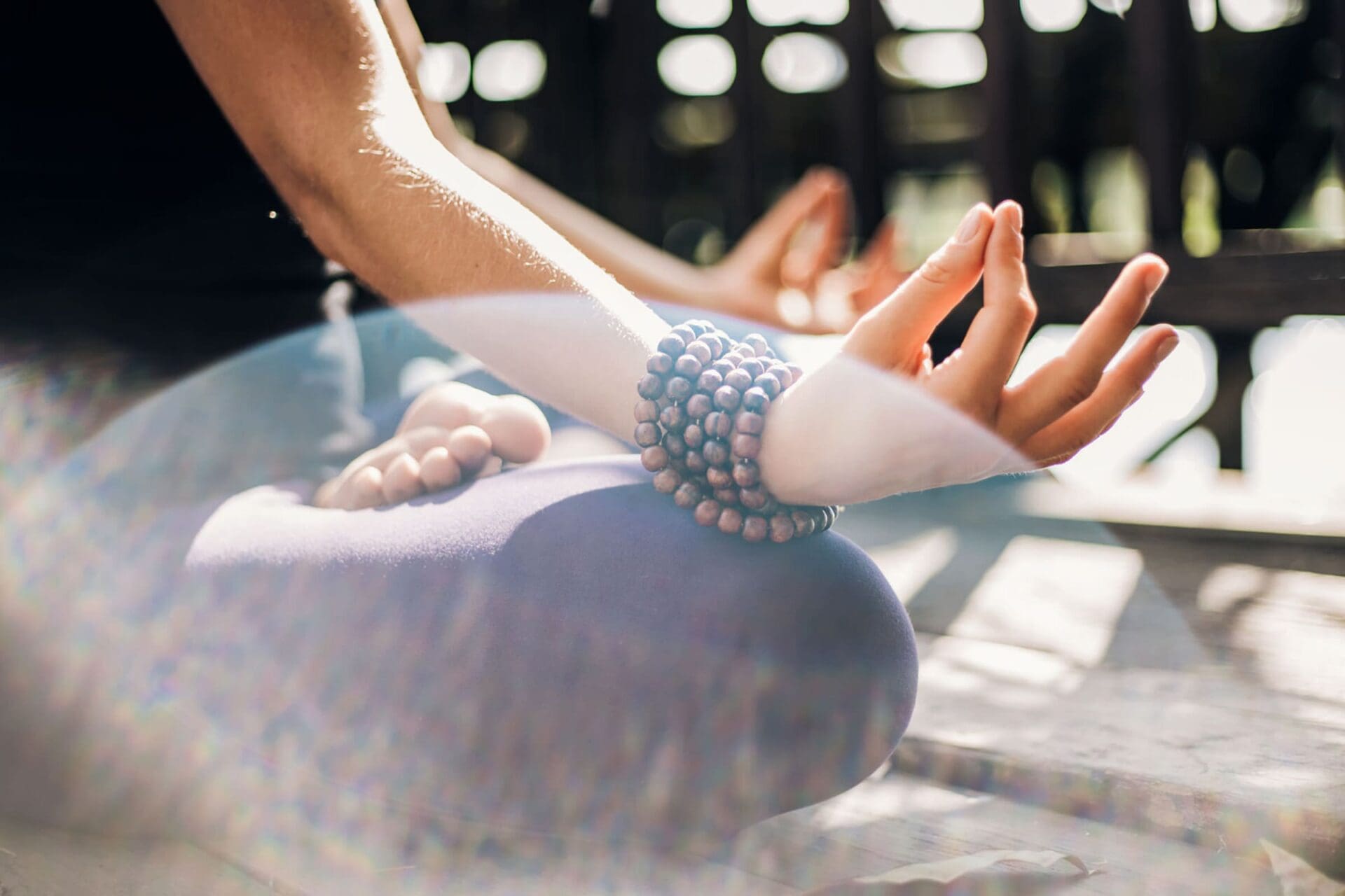 A person sits cross-legged on the ground, meditating with one hand resting on a knee in a mudra pose, wearing beaded bracelets. Sunlight and lens flares create a peaceful, serene atmosphere.