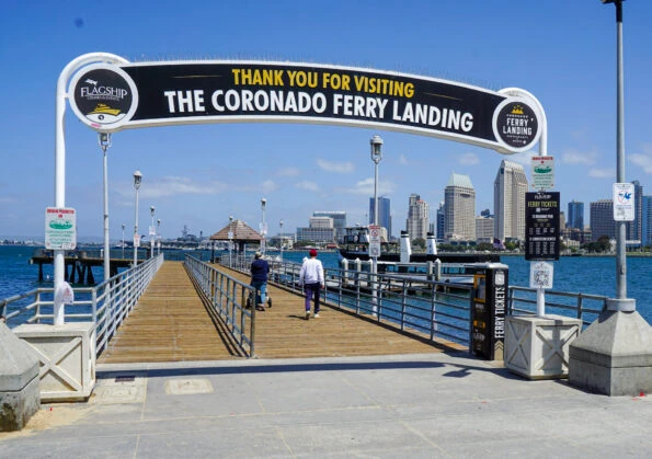 Entrance to the Coronado Ferry Landing with a sign overhead, a wooden pier extending over the water, two people walking on the pier, and the San Diego city skyline in the background under a clear sky.