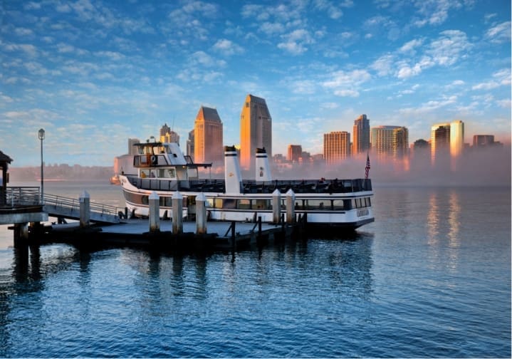 A white ferry boat is docked at a pier, with calm water in the foreground and a city skyline, including tall buildings, visible across the bay under a partly cloudy sky at sunrise or sunset.