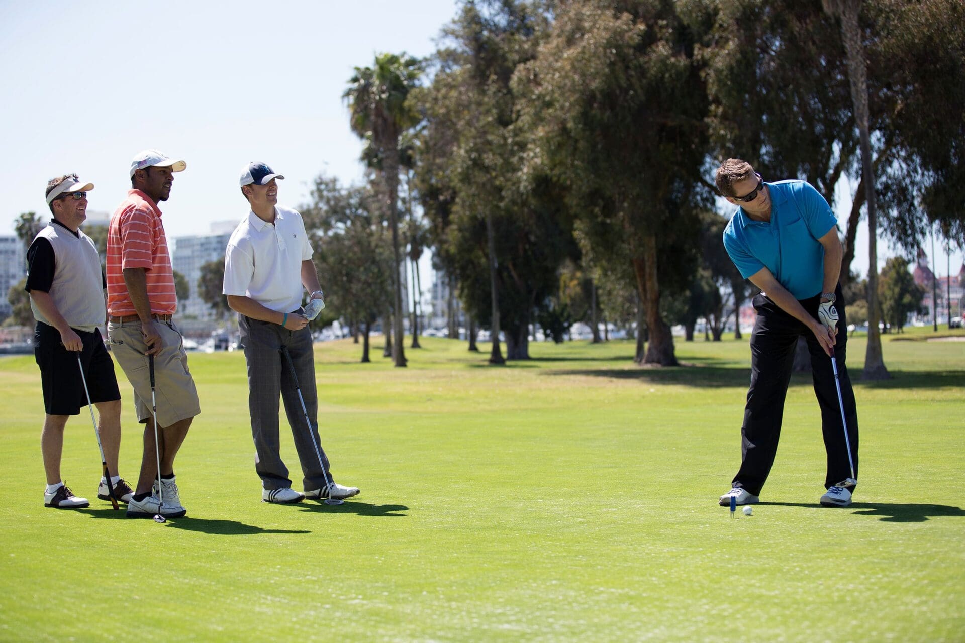 Four men are on a sunny golf course. Three watch as the fourth, wearing a blue shirt, prepares to putt the golf ball on the green. Trees and buildings are visible in the background.
