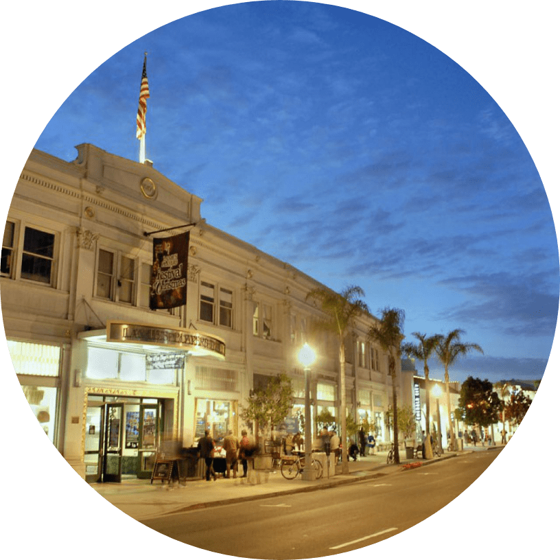 A historic white building with an American flag on top, lit by streetlights at dusk. People are sitting outside a restaurant on a palm-lined street under a blue evening sky.