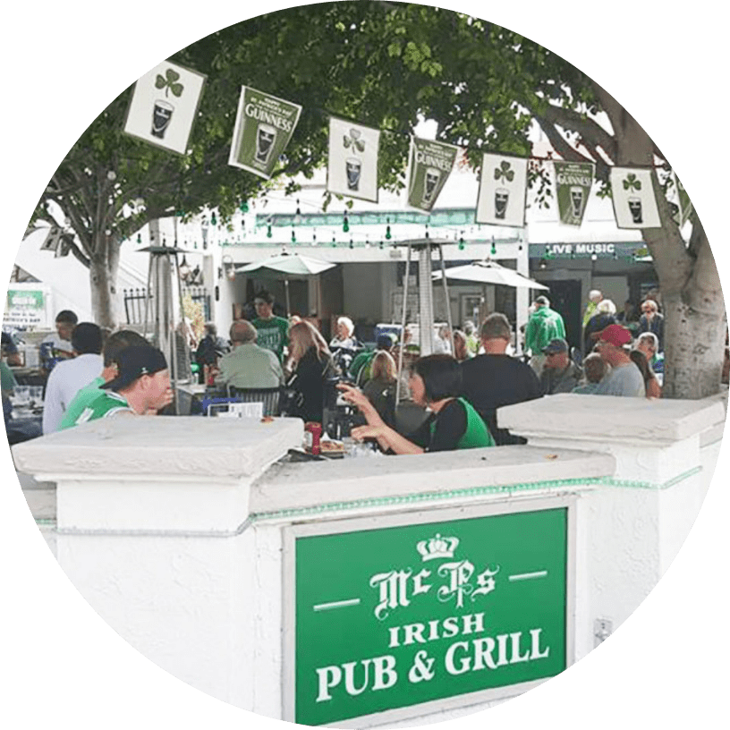 People enjoy food and drinks on the outdoor patio of McPs Irish Pub & Grill, decorated with shamrock banners and Guinness signs, under leafy trees. The pub is busy and festive, with green attire visible.