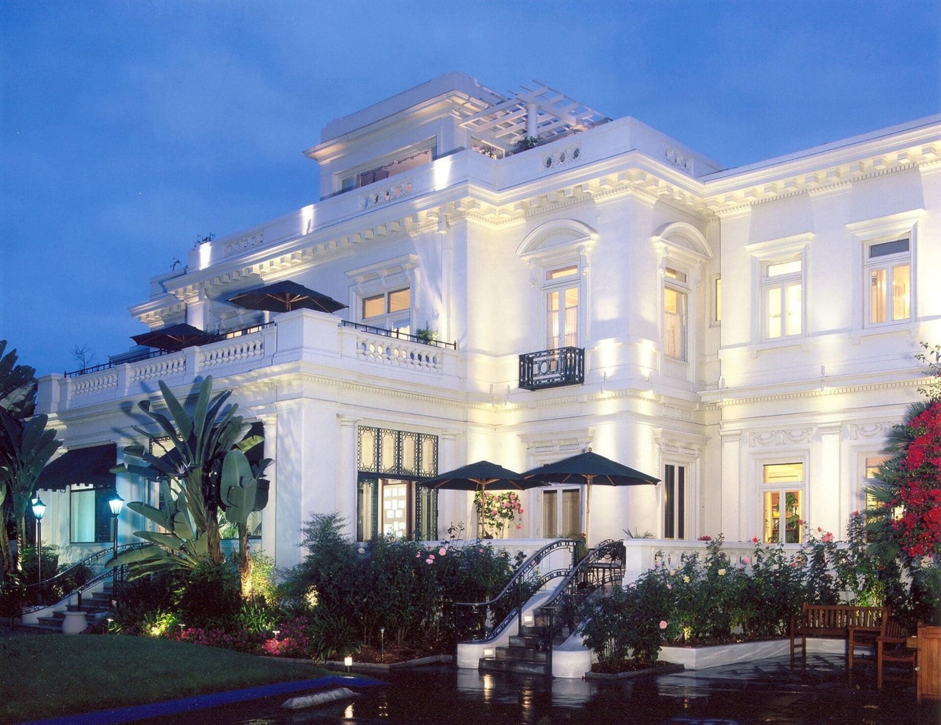 A large, elegant white mansion with ornate architectural details, glowing with exterior lights at dusk; surrounded by lush gardens, palm trees, umbrellas, and a wet stone path reflecting the lights.