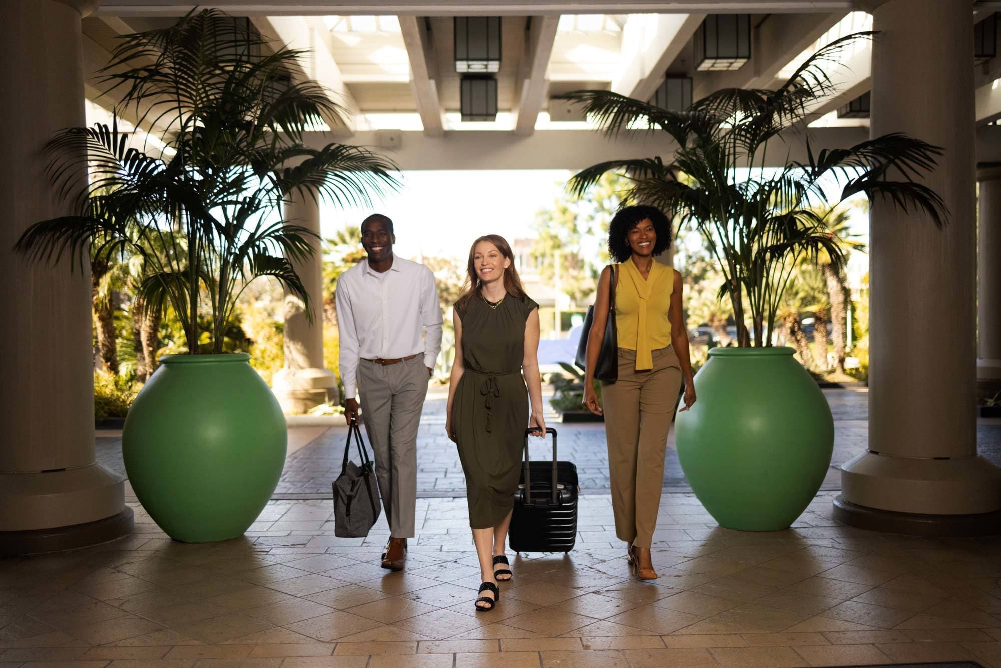 Three people walk through a bright, modern lobby with large green planters, smiling and carrying luggage and bags, suggesting they are traveling or arriving at a hotel.