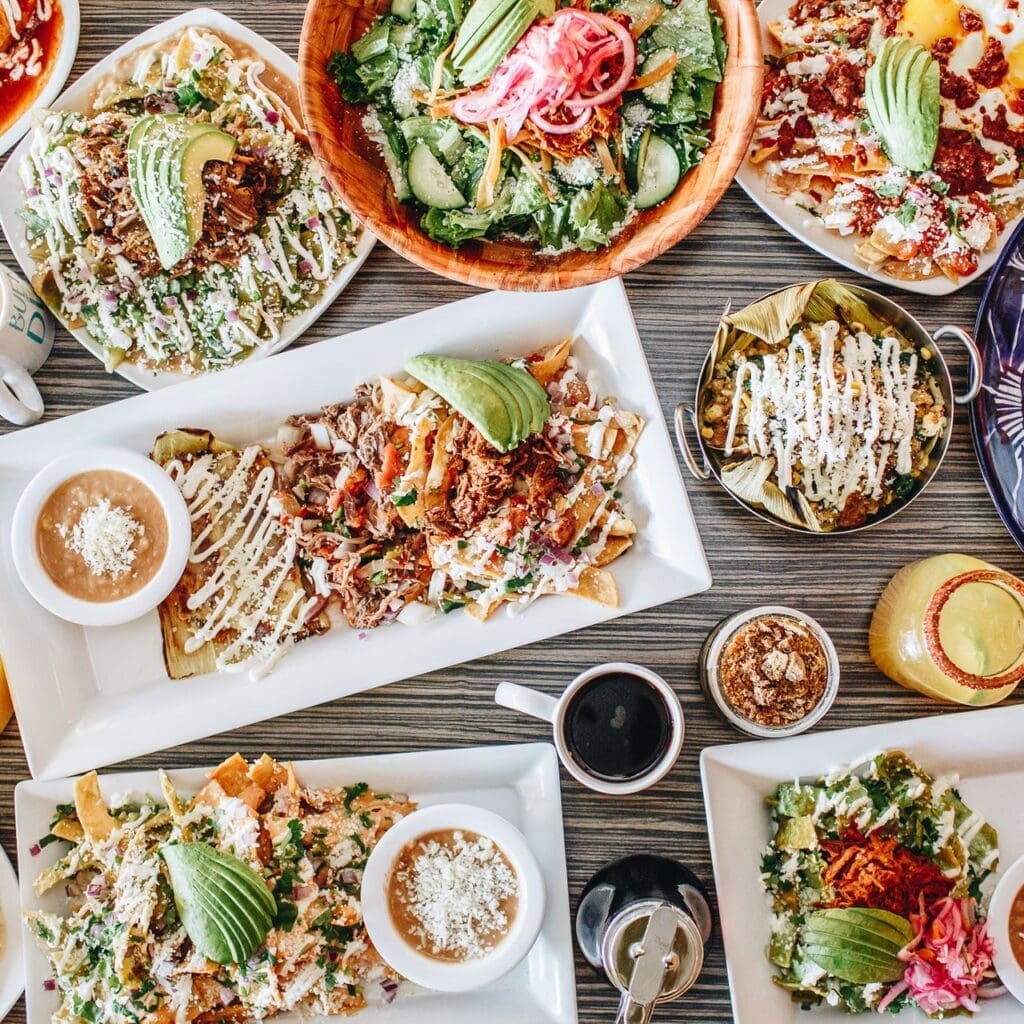 A tabletop filled with colorful Mexican dishes, including nachos, salads, and enchiladas, all topped with avocado, sauces, and cheese. Drinks and condiments are also arranged around the plates.
