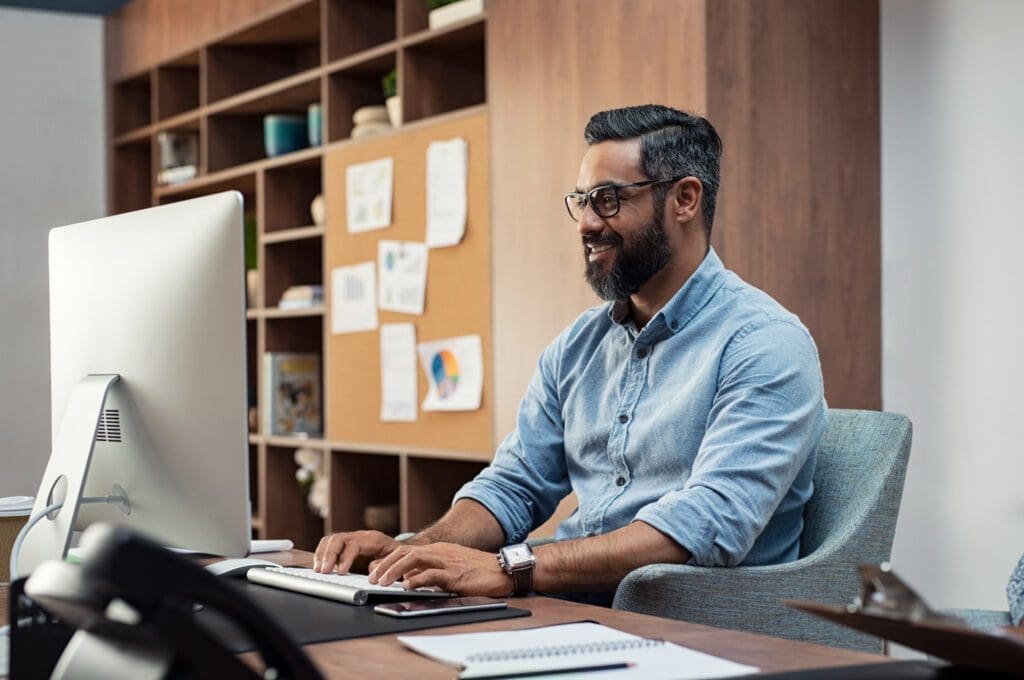 A man with a beard and glasses sits at a desk, smiling while working on a computer in a modern office with shelves and a corkboard in the background.