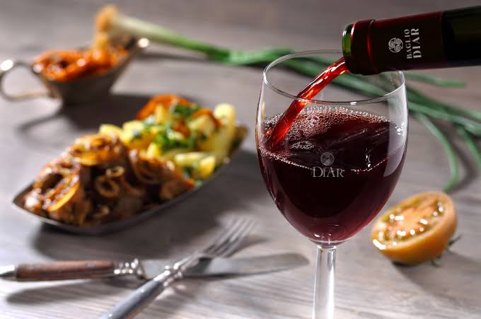 Red wine is being poured into a glass, with plated food, a fork and knife, and a sliced lemon in the background on a wooden table.