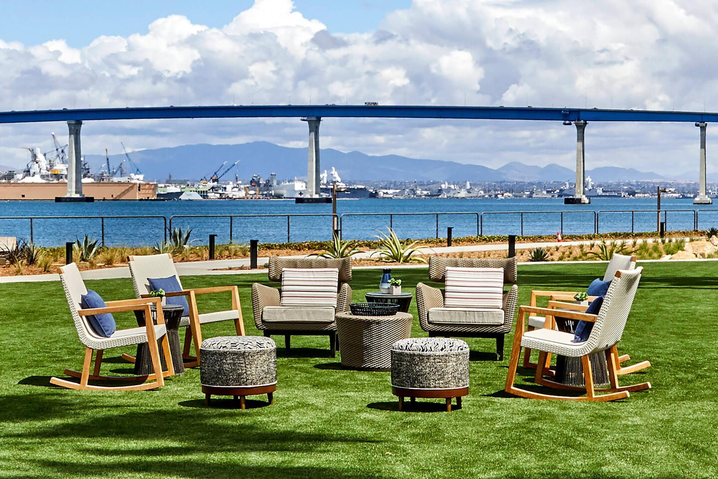 Outdoor seating area with cushioned chairs and ottomans arranged in a circle on green grass, overlooking water with ships and a tall blue bridge in the background, mountains and clouds in the distance.